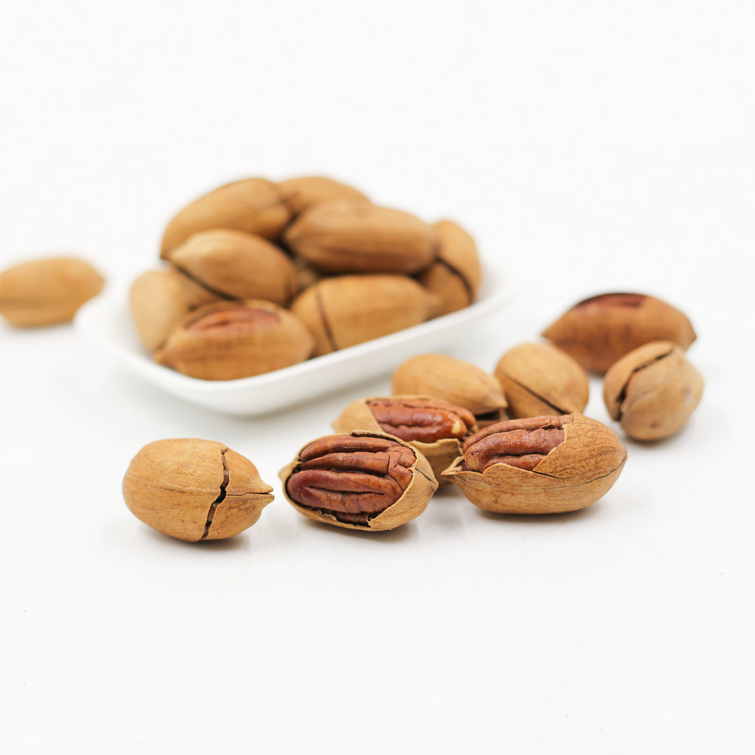 Several pecans and a bowl of whole pecans arranged on a clean white background, with some nuts cracked open to show the brown nut interior.